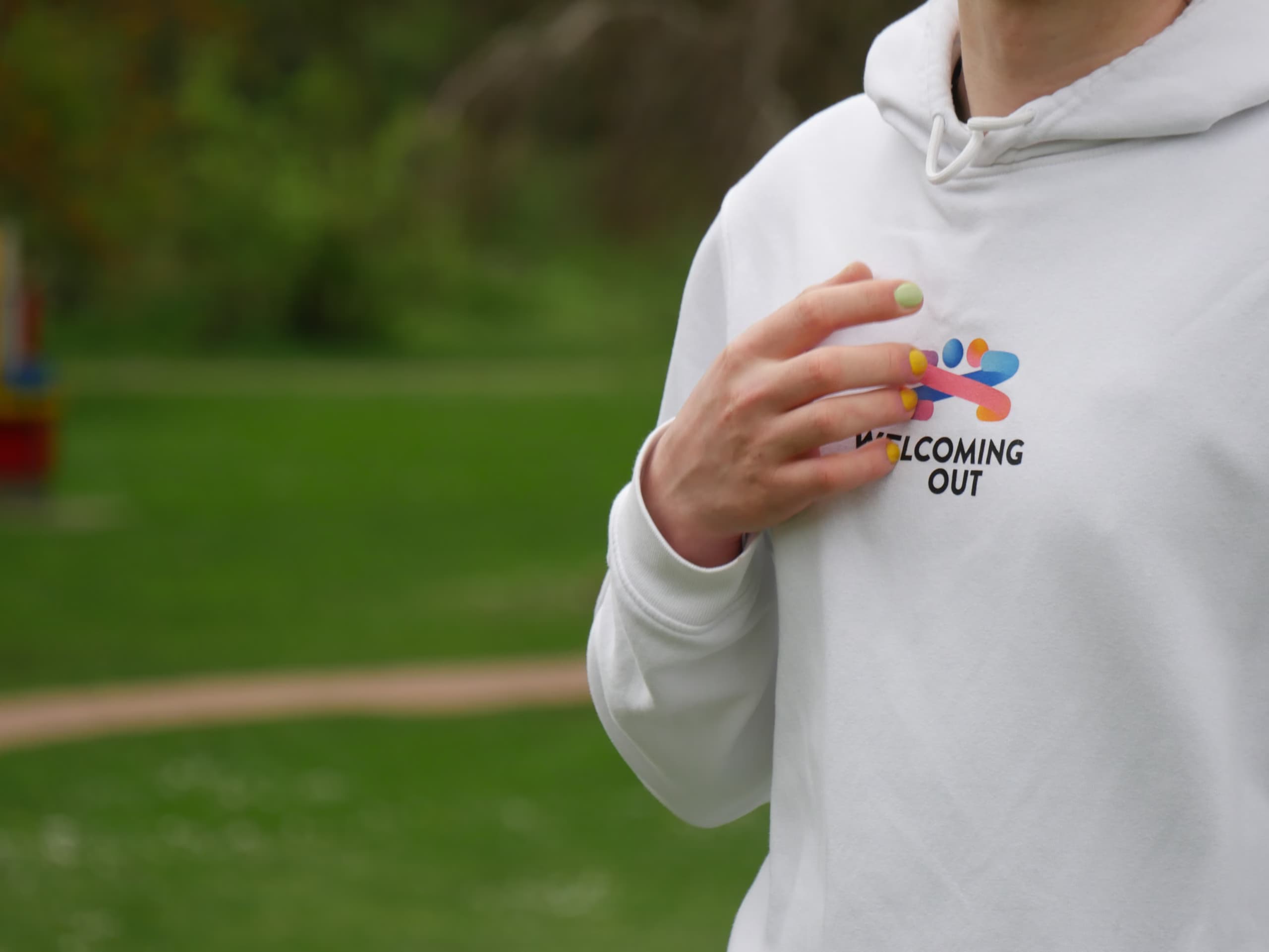 Person wearing a white hoodie with a colorful logo and "Welcoming Out" text, standing in a park. They have nails painted in various colors.