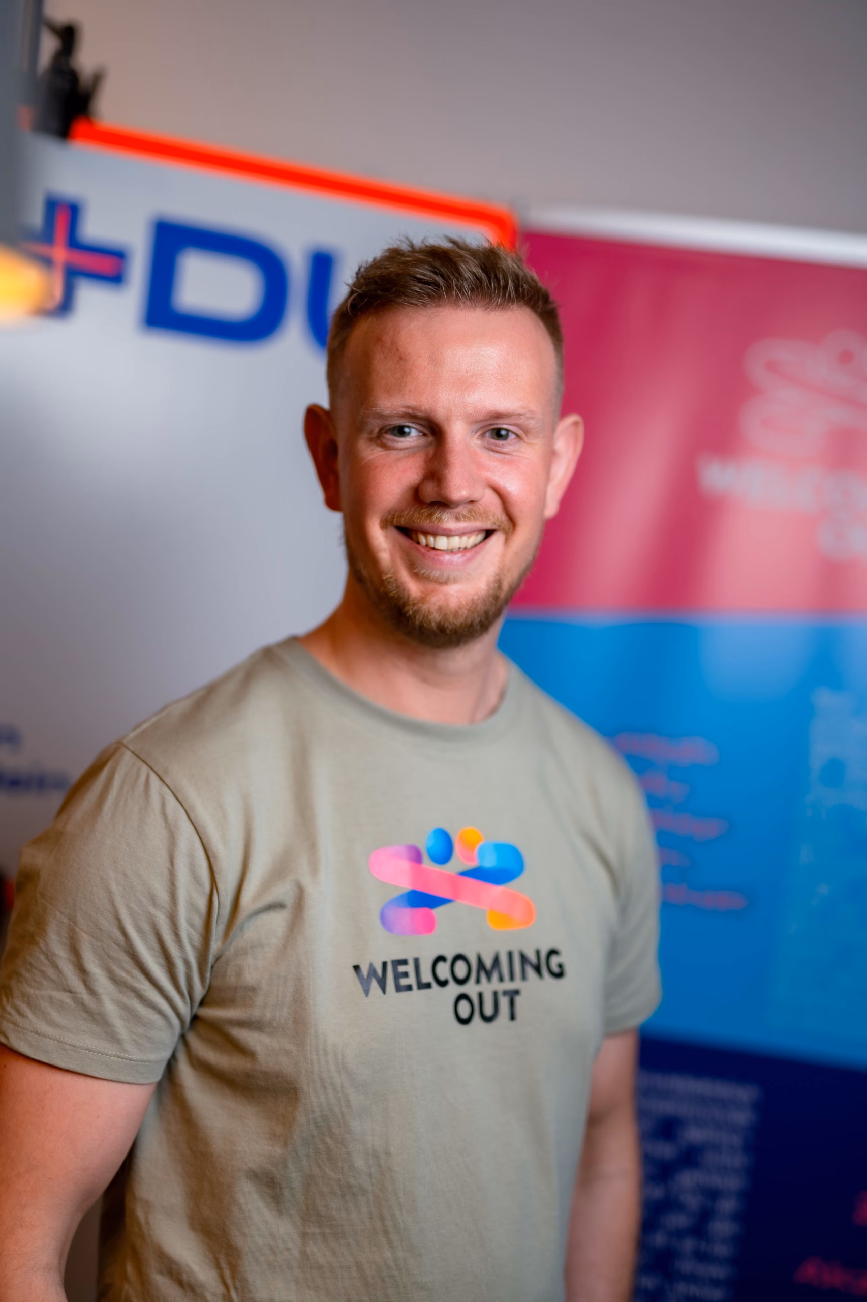 Progetto senza titolo (12) Person wearing a white hoodie with a colorful logo and "Welcoming Out" text, standing in a park. They have nails painted in various colors.