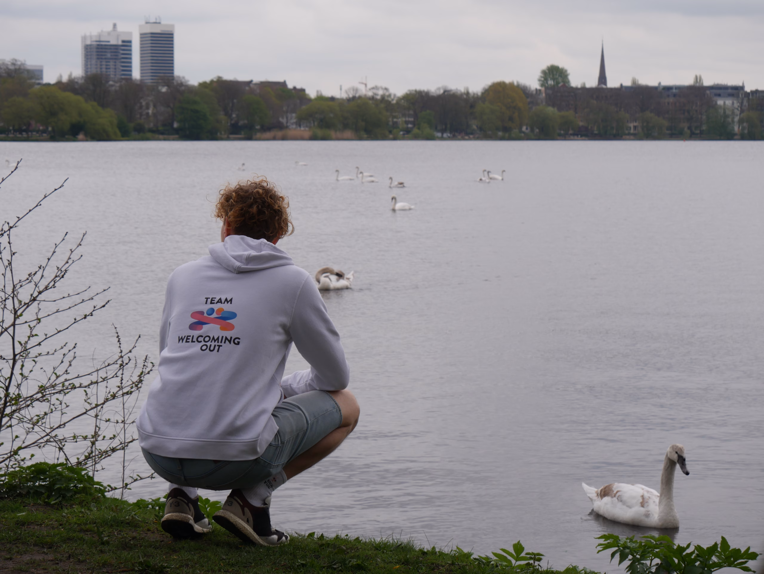 Progetto senza titolo (12) Person wearing a white hoodie with a colorful logo and "Welcoming Out" text, standing in a park. They have nails painted in various colors.